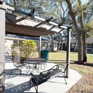 A backyard patio featuring a pergola with seating and a table surrounded by trees.