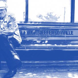 A monochrome image with a blue tint showing a man sitting on a bench, possibly waiting.