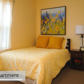 A cozy bedroom with a bed covered in yellow bedding, a floral painting, and a lamp on the nightstand.