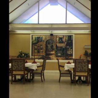 A well-lit dining room featuring tables and chairs, with a large artwork on the wall and a high ceiling with windows.