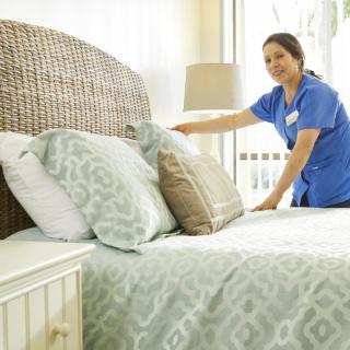 A housekeeper in a blue uniform making a bed in a bright and well-decorated bedroom.