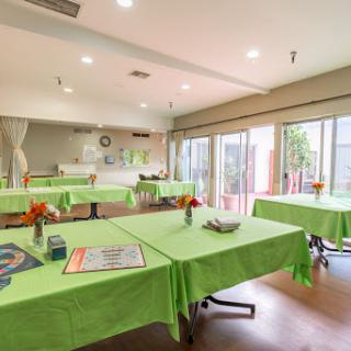 A spacious event room set up with green tablecloths and floral centerpieces, featuring ample natural light.