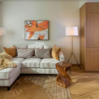 A cozy living room with a white sectional sofa, adorned with cushions, and an abstract painting on the wall. A floor lamp adds warmth to the space, complemented by a wooden side table and a textured rug.