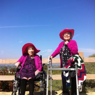 Two elderly women enjoying a sunny day outdoors, wearing matching red hats and pink outfits. One is in a wheelchair and the other uses a walker.