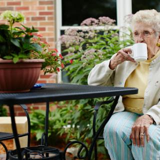 An elderly woman enjoys a cup of tea or coffee while sitting in a serene outdoor setting surrounded by plants.