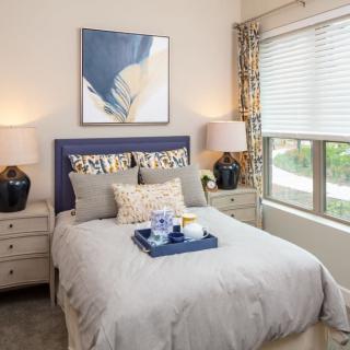 A well-decorated bedroom featuring a neatly made bed with decorative pillows, flanked by two nightstands with lamps, and a piece of abstract artwork above the headboard. Large windows allow natural light to illuminate the room.