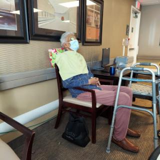 An elderly man wearing a mask sits in a waiting area, accompanied by a walker.