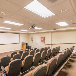 An empty conference room with rows of chairs facing a large screen, equipped with a projector.