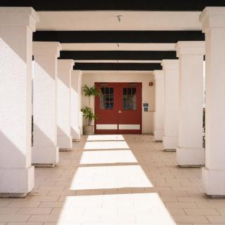 Sunlight casts shadows along a columned corridor leading to a red door.