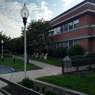 A view of a brick building surrounded by green landscaping, featuring a pathway and seating area.