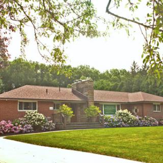A charming single-story brick house surrounded by lush greenery and beautifully landscaped flowers.