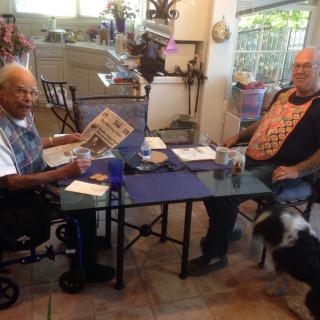 Two individuals are seated at a table indoors, with one reading a newspaper. A dog is nearby.
