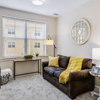 A cozy living room with a sofa, decorative pillows, and a large window, featuring a mirror and warm lighting.