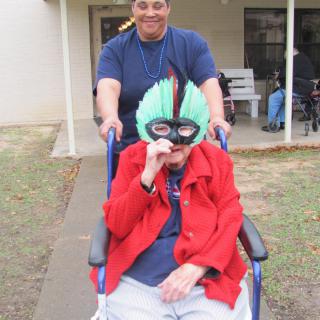 An elderly person wearing a colorful mask is being pushed in a wheelchair by a caregiver outside.