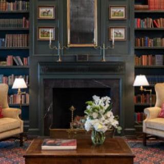 A cozy living room featuring a library with dark green walls, elegant armchairs, and a warm fireplace.