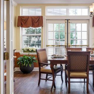 A well-lit dining room with a wooden table and chairs, large windows, and indoor plants.