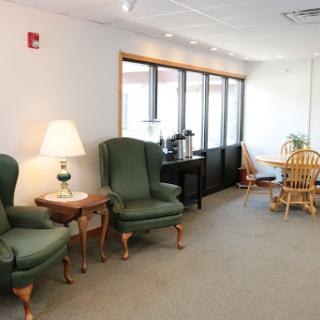 A cozy waiting area featuring comfortable green chairs, a lamp, and a small dining table bathed in natural light.