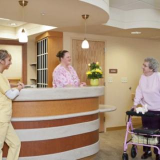 Two nurses interact with an elderly woman using a walker at a hospital reception area.