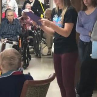 A caregiver reads to a group of elderly individuals in a nursing home setting.