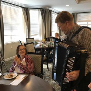 A musician plays the accordion for an elderly woman in a dining room setting.