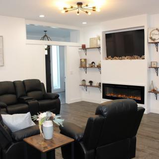A modern living room with a cozy arrangement of black leather sofas and armchairs, featuring a wall-mounted fireplace and decorative shelves.