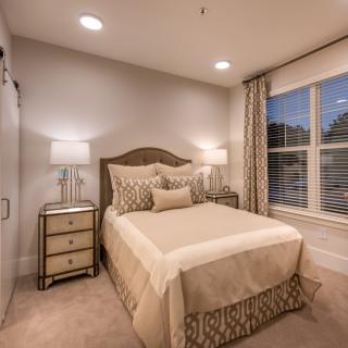 A cozy and elegantly designed bedroom with neutral tones, featuring a neatly arranged bed, bedside tables with lamps, and large windows with drapes.