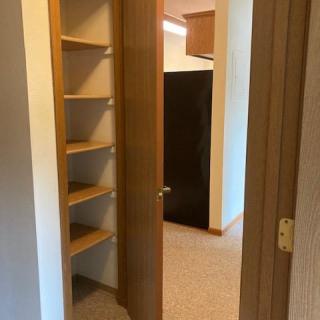 A hallway with a closet featuring wooden shelves and an open door leading to another room.