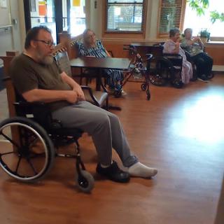 An elderly man seated in a wheelchair, surrounded by other seniors in a care facility.
