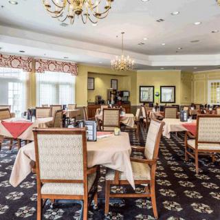 A spacious and elegantly decorated restaurant dining room with multiple tables and chairs, featuring chandeliers and carpeted flooring.
