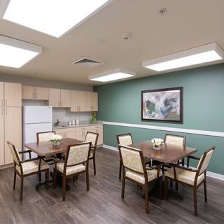 A cozy dining area with a kitchen, featuring wooden tables and chairs, green walls, and overhead lighting.