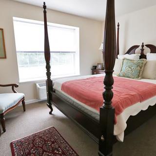 A cozy bedroom featuring a four-poster bed with a red blanket, an upholstered chair, and a window providing natural light.