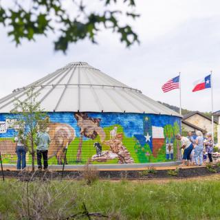 A mural painted on a silo with people gathered around, accompanied by American and Texas flags.