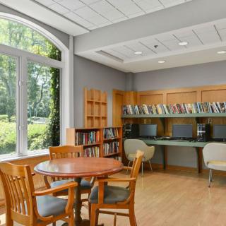 A cozy library corner featuring a reading table, bookshelves, and computer stations by large windows.