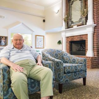An elderly man sitting comfortably in a living room with cozy furniture and a fireplace.