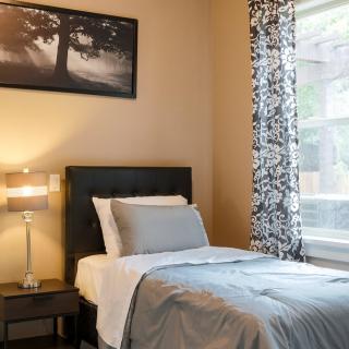 A cozy bedroom with a neatly made bed, a bedside lamp, and a window with patterned curtains.