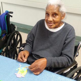 An elderly woman in a wheelchair engaged in a crafting activity at a table.