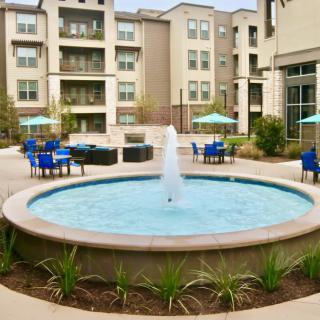 A modern apartment complex courtyard featuring a central fountain and outdoor seating with patio umbrellas.