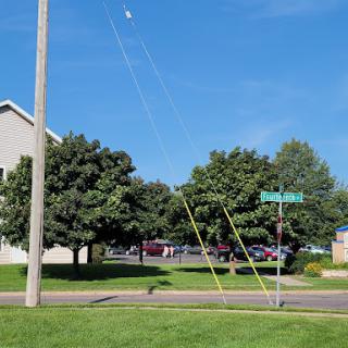 A utility pole stands alongside a road, with multiple wires extending towards the clear blue sky and trees in the background.