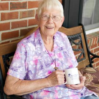 An elderly woman smiling while sitting on a bench and holding a mug.