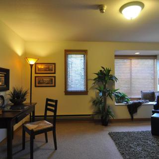 A cozy living room with warm lighting, featuring a desk and chair, a window seat, and decorative plants.