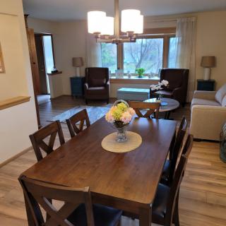 A cozy dining and living room area with a wooden table, chairs, and natural lighting from large windows.