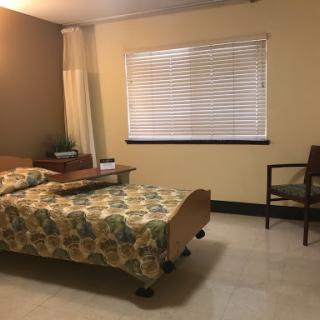 A simple and cozy bedroom featuring a bed with floral bedding, a window with blinds, and a wooden chair.