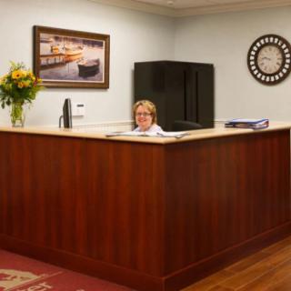 A woman sits at a reception desk in an office, with flowers, a clock, and a painting on the wall.