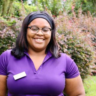 A woman in a purple shirt smiling outdoors with greenery in the background.