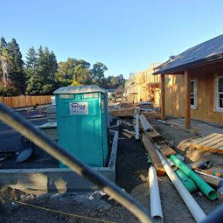 A view of a construction site featuring unfinished buildings, pipework, and a portable toilet under clear skies.