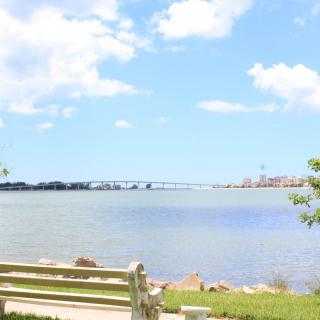 A serene view of a bench by the water with a bridge in the background under a bright, cloudy sky.