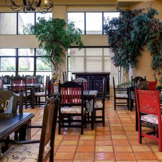 A cozy restaurant dining area with wooden tables and chairs, tiled flooring, large windows, and decorative plants.