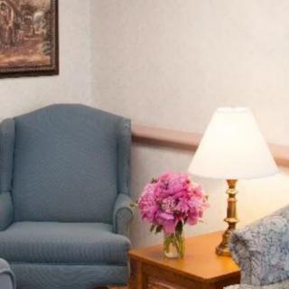 A cozy living room corner with a blue armchair, a lamp on a wooden table, and a bouquet of pink flowers.