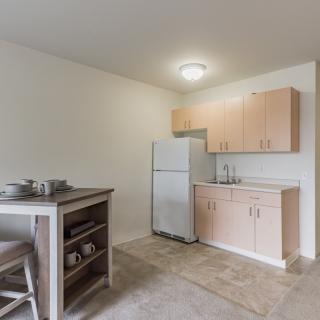 A minimalist kitchen with wooden cabinets, a white refrigerator, and a small dining table with stools.