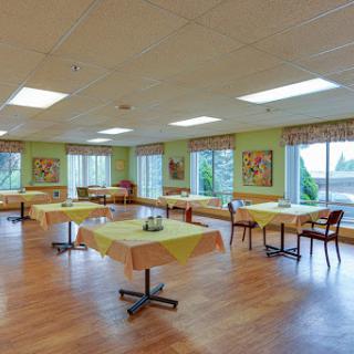 A spacious dining room featuring several tables and chairs, adorned with green walls and artwork, illuminated by natural light from large windows.